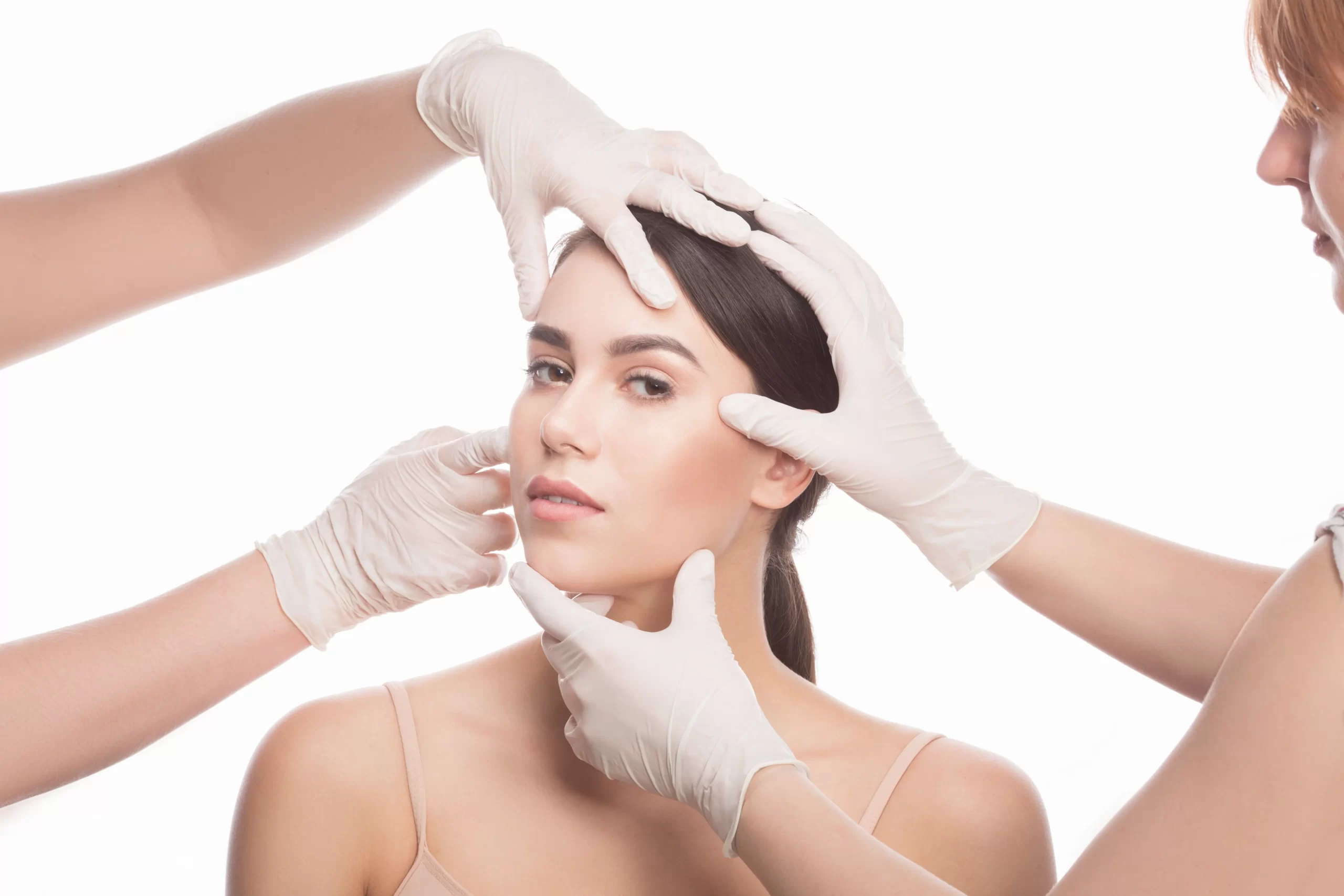 handmade-beauty-beautiful-young-woman-keeping-eyes-opened-while-four-hands-medical-gloves-touching-her-face-isolated-white-background
